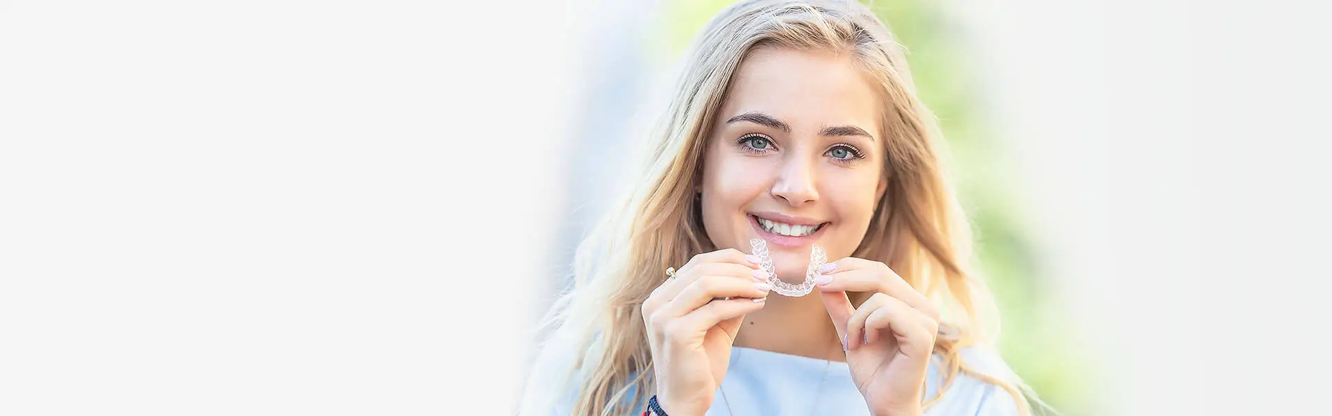 suresmiledental-invisalignheader-1 A smiling woman with long blonde hair holds a clear dental aligner in her hands. She is outdoors, wearing a light blue top, against a blurred background.